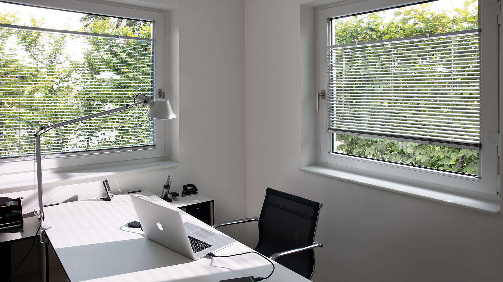 Homeoffice mit zwei Fenstern und silberfarbenen Jalousien. Lamellen regulieren das Licht für blendfreies Arbeiten.